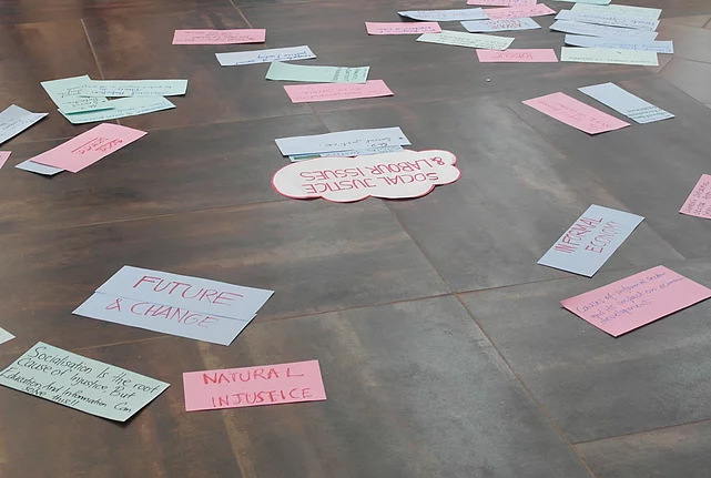Scattered across the floor are various colourful cards featuring terms and ideas such as ‘Informal Economy’, ‘Future & Change’ and ‘Natural Injustice’.